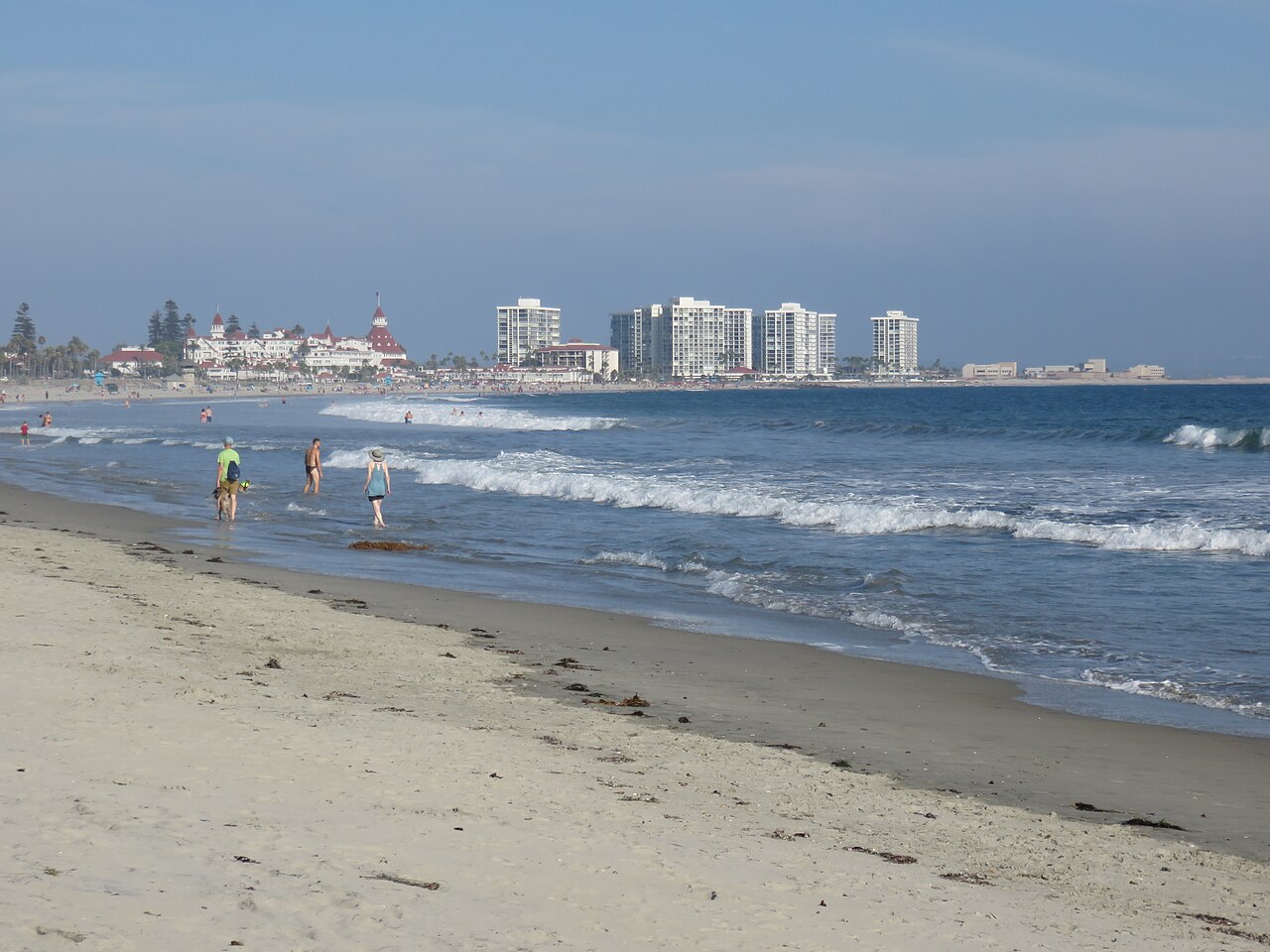 A wide sandy beach with small waves, the red-roofed Hotel del Coronado, and white towers down the shore.