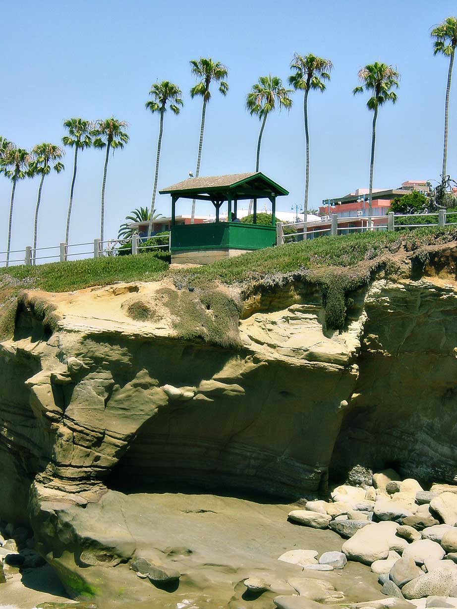 Sandstone caves under a green wooden shelter and a row of palm trees at La Jolla Cove.