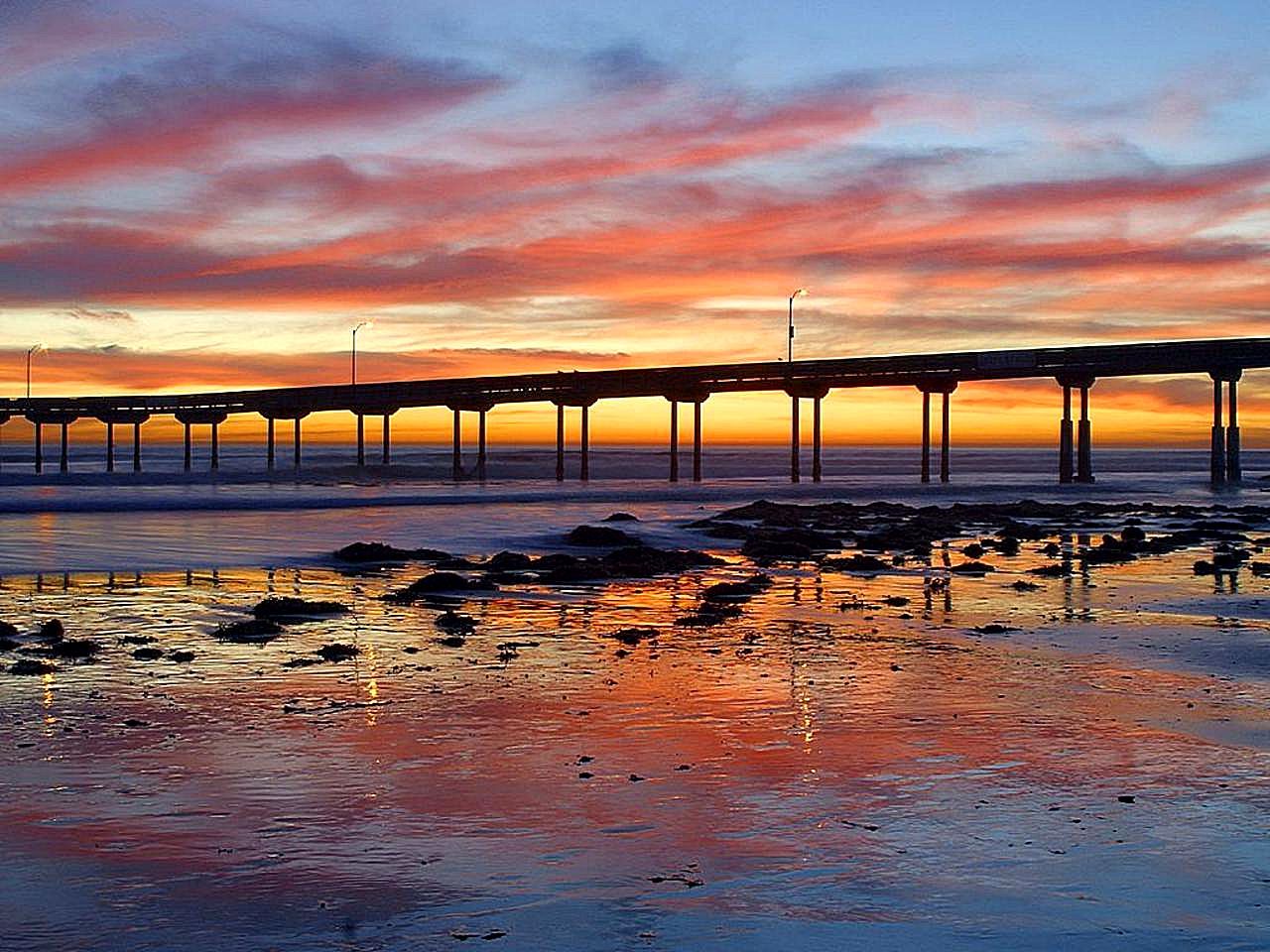 The Ocean Beach Pier at sunset, a long concrete pier on pilings over wet sand and rocks.