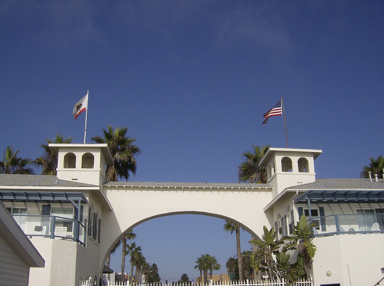 The white cottage arch on Crystal Pier, with a California flag and an American flag, palm trees behind it.